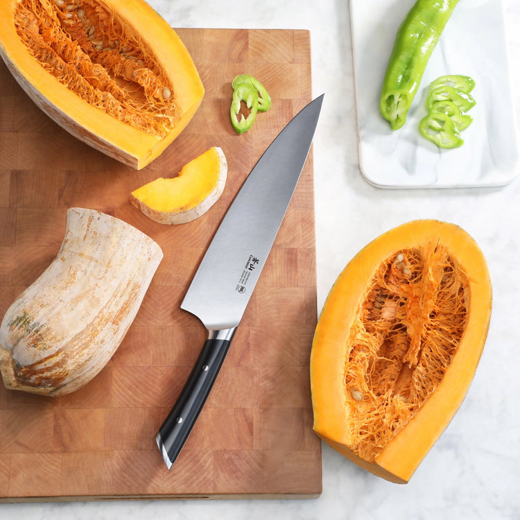 10-inch chef's knife with black handle on wooden cutting board next to halved pumpkin and sliced green pepper.