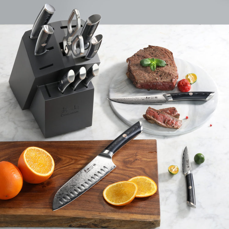 12-piece knife set with black handles in a block, displayed with sliced oranges, steak, and a steak knife on a marble surface