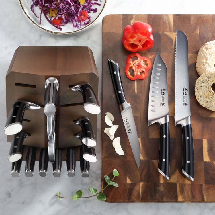 Wooden knife block with six black-handled knives next to a cutting board holding three knives, sliced red bell pepper, garlic