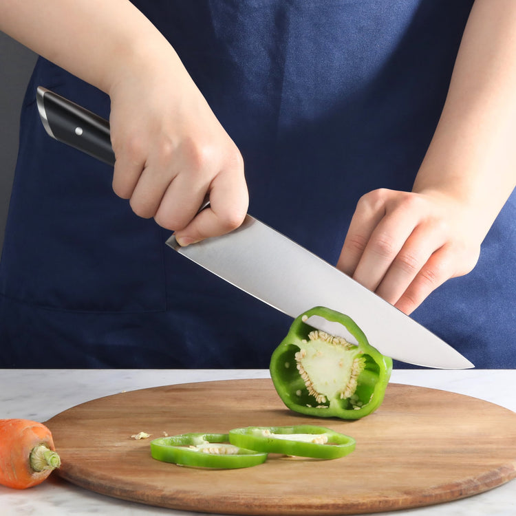 Hands slicing a green bell pepper on a round wooden cutting board with a black-handled chef knife.