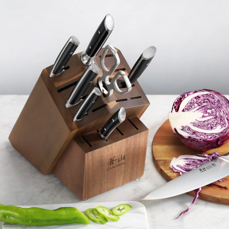 Wooden knife block with six knives and kitchen shears, placed on a countertop near sliced vegetables and a cutting board.