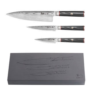 
                    
                      Three Damascus steel knives with black handles and red accents displayed above a gray box engraved with knife outlines.
                    
                  
