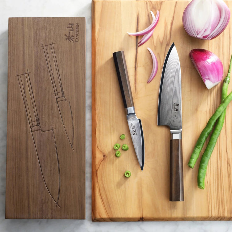 Two Damascus steel knives with wooden handles on a wooden cutting board next to sliced onion and green chili, with a wooden b