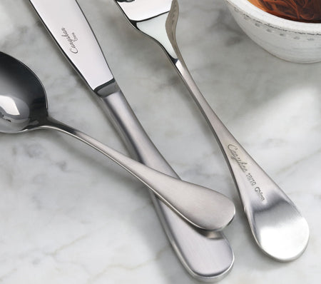 Three stainless steel flatware pieces with matte finish handles on a marble surface near a bowl.