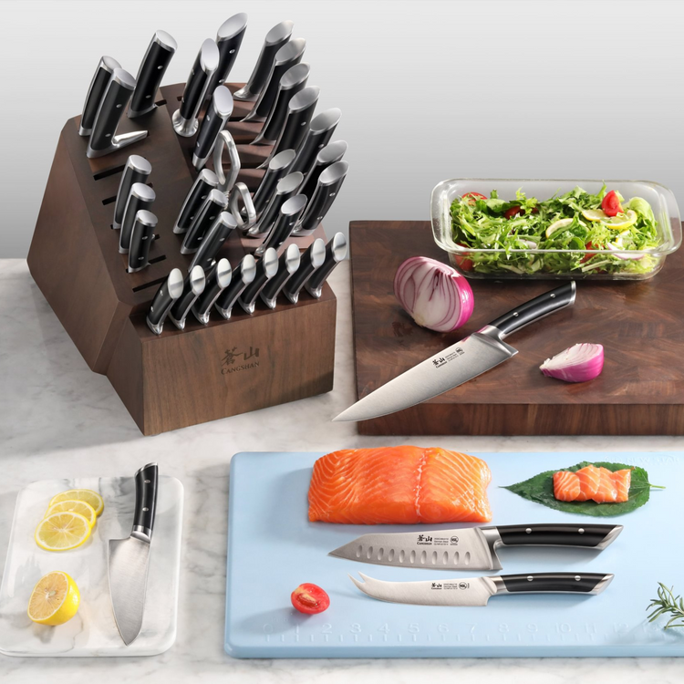 Wooden knife block set with multiple black-handled knives, cutting boards with salmon, onion, lemon, and a glass dish of sala