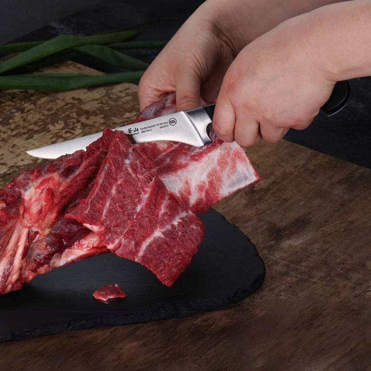 Hands slicing raw beef ribs with a knife on a black cutting board and wooden surface.