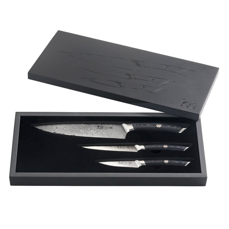 Three knives with patterned blades and black handles arranged inside an open black wooden box with a matching lid.