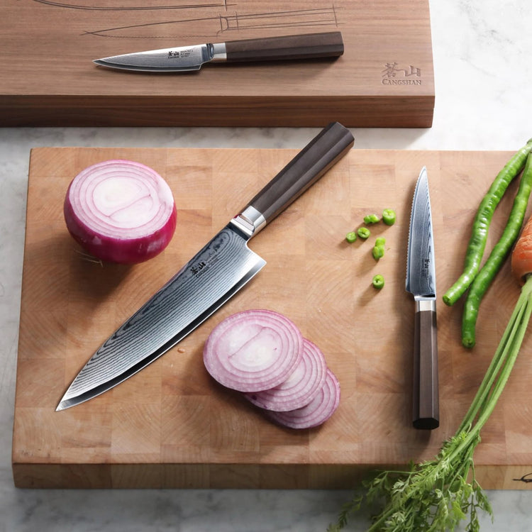 Three knives with dark wooden handles on wooden cutting boards, one slicing a red onion, surrounded by vegetables.