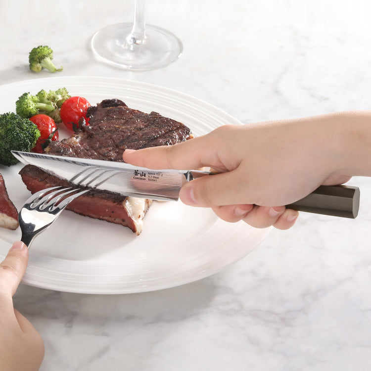 Hand holding a steak knife cutting a medium-rare steak on a white plate with broccoli and cherry tomatoes.