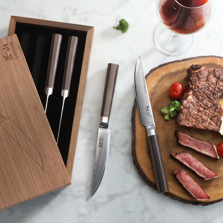 Four steak knives with dark handles in a wooden box, two knives beside a sliced steak on a wooden board with cherry tomatoes