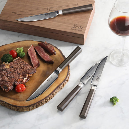 Four steak knives with dark handles and Damascus steel blades, wooden box, sliced steak on wooden board, glass of red wine.
