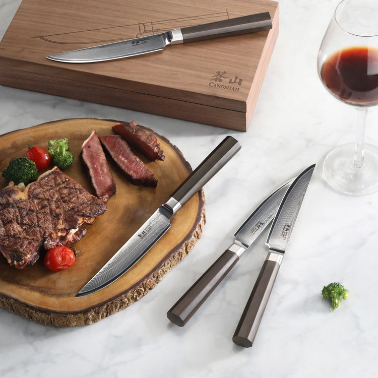 Four steak knives with dark handles and Damascus steel blades, wooden box, sliced steak on wooden board, glass of red wine.