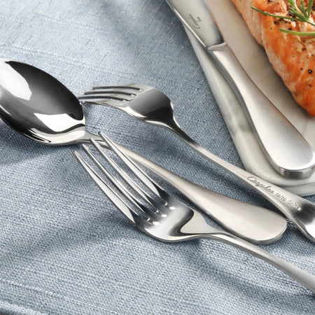 Stainless steel flatware with matte finish handles arranged on a blue cloth next to a plate with cooked salmon.