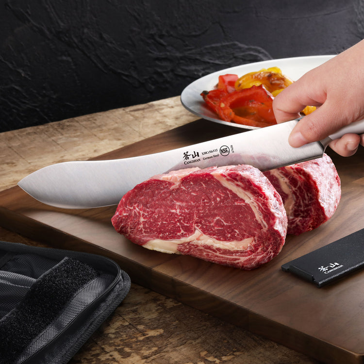 Hand holding a large knife slicing raw marbled steak on a wooden cutting board with grilled vegetables in the background.