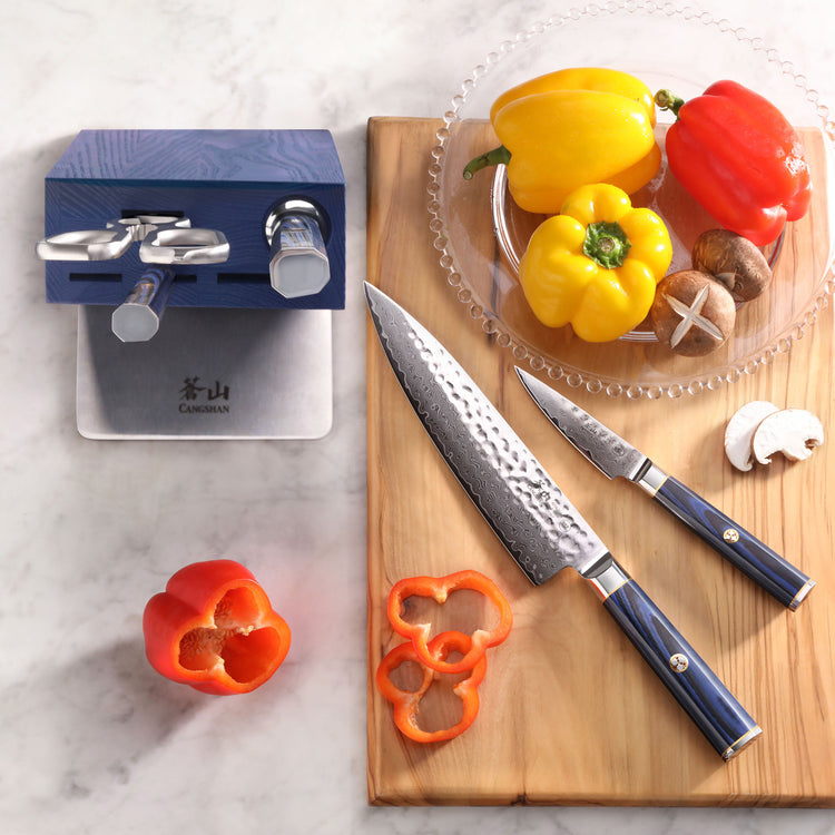 Two knives with blue handles on a wooden cutting board next to sliced red bell pepper, whole peppers, and mushrooms, with a b