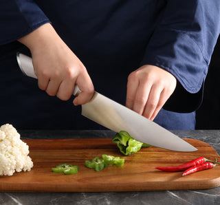
                    
                      Hands slicing a green bell pepper on a wooden cutting board with cauliflower and red chili peppers nearby.
                    
                  