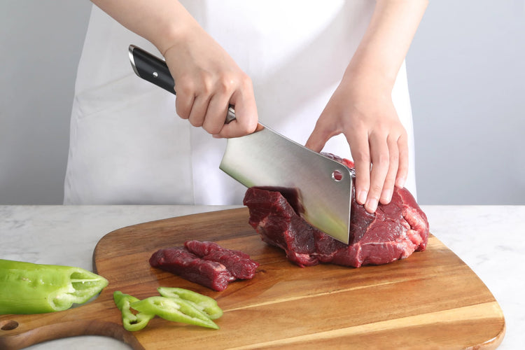 Person slicing raw red meat on wooden cutting board with a large black-handled cleaver knife.
