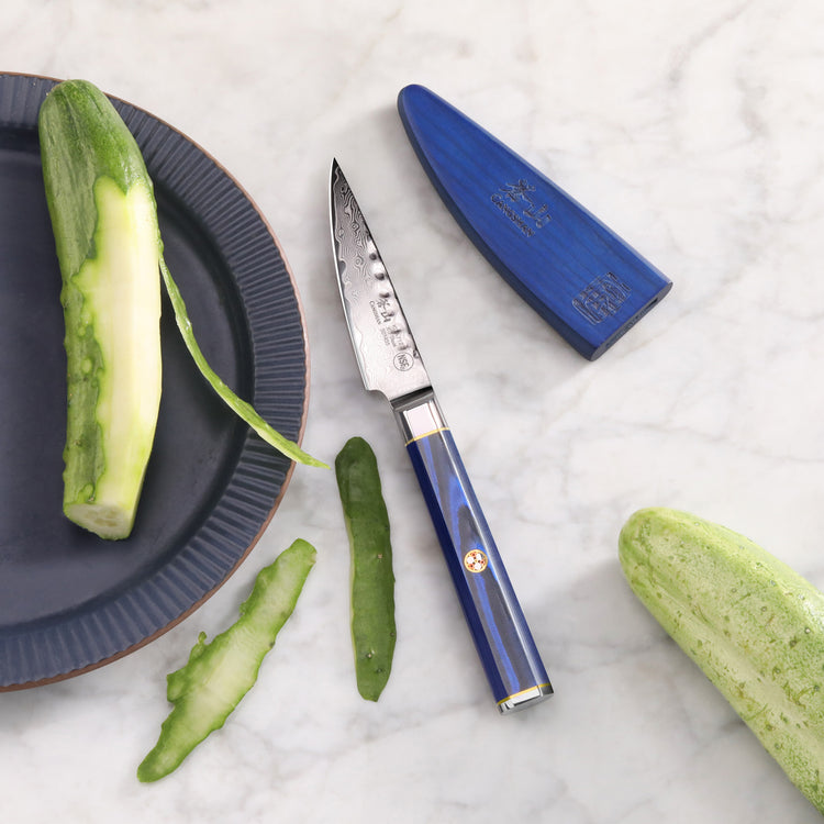 3.5-inch paring knife with blue handle and sheath, partially peeled cucumber on plate and marble surface.