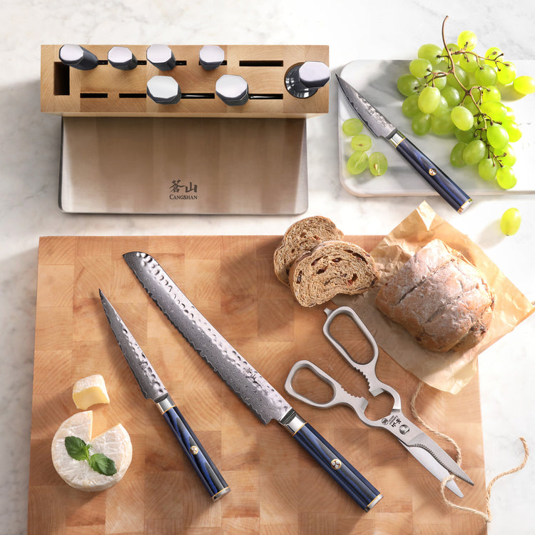 Knife block with seven knives, two blue-handled knives, a bread knife, a cheese knife, sliced bread, grapes, and cheese on a