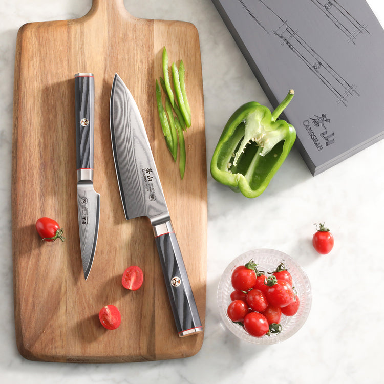 Two Damascus steel knives with black handles on a wooden cutting board, surrounded by sliced green peppers and cherry tomatoe