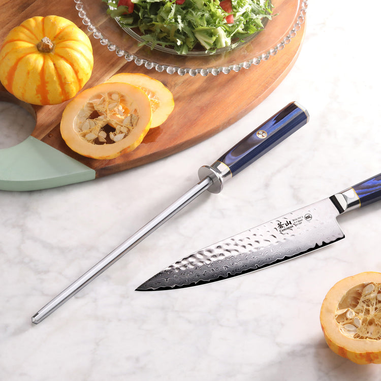 Blue-handled honing steel and chef's knife on marble countertop with sliced squash and salad on wooden board.