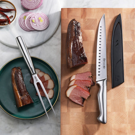 Two-piece carving set with a fork and a serrated knife with sheath, displayed with sliced meat and onion rings.