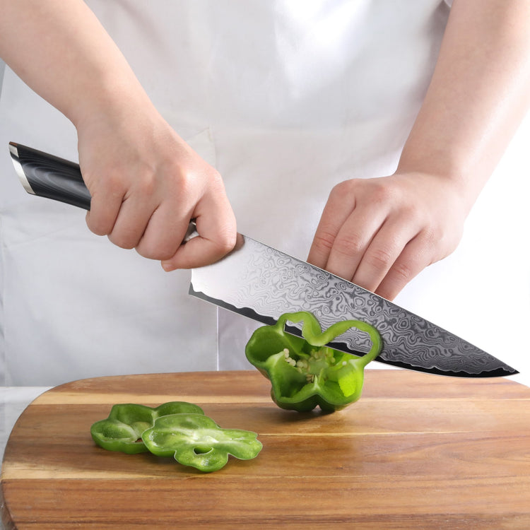 Hands holding a Damascus steel knife slicing a green bell pepper on a wooden cutting board.