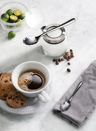 
                    
                      Polished stainless steel sugar spoon resting on a sugar jar lid, with coffee cup, cookies, and coffee beans on a marble surfa
                    
                  