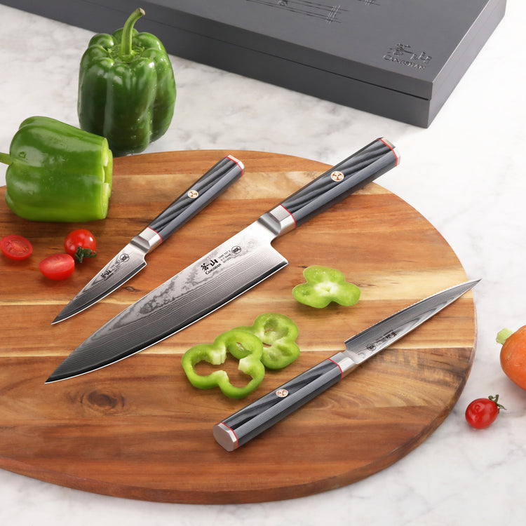 Three kitchen knives with black handles and Damascus steel blades on a round wooden cutting board with sliced green bell pepp