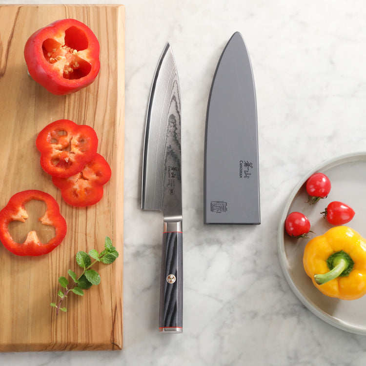 YARI Series 8-Inch Chef's Knife with sheath on marble surface, next to sliced red bell pepper on wooden board and yellow bell