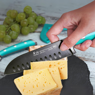 
                    
                      Hand holding a turquoise-handled knife slicing semi-hard cheese on a slate board with green grapes in the background.
                    
                  