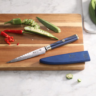 
                    
                      Serrated utility knife with blue handle and sheath on wooden cutting board with sliced okra and red chili peppers.
                    
                  