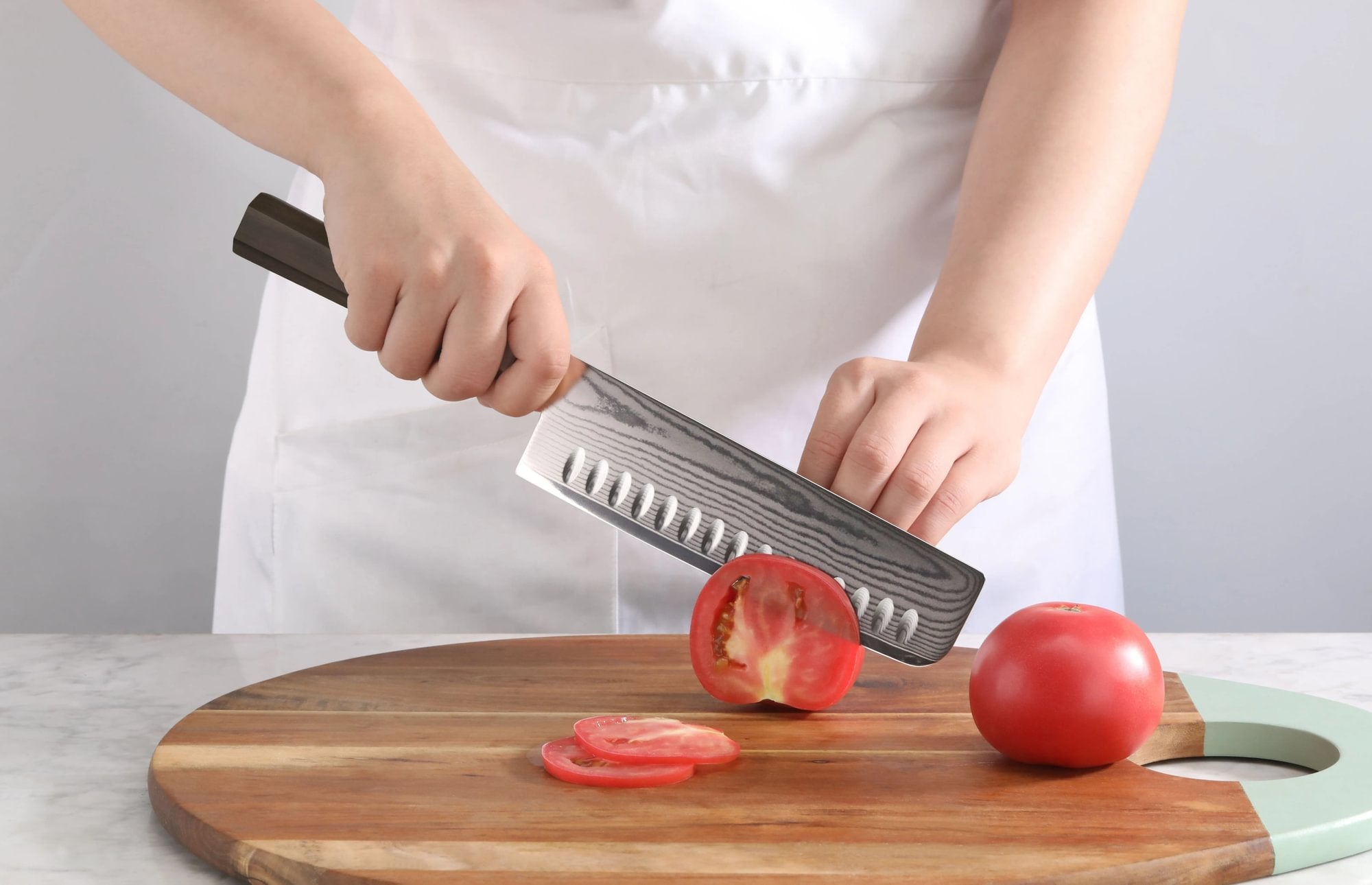 A person slicing a red tomato on a wooden cutting board using a MAYA Series 7-Inch Nakiri Knife.