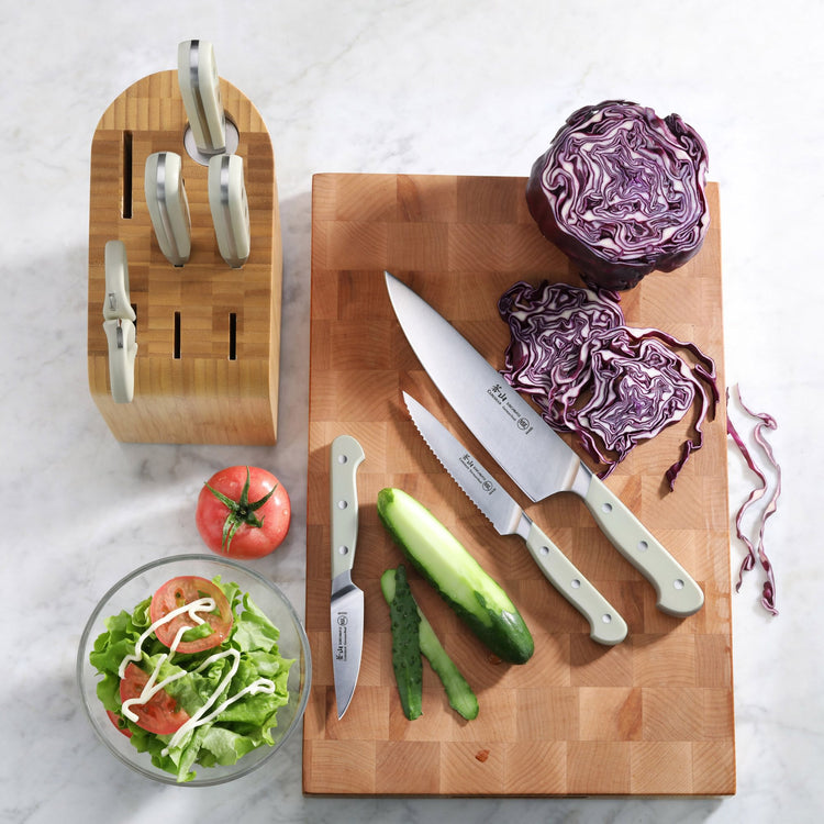 Bamboo knife block with four knives, three knives on cutting board with sliced cucumber and red cabbage, salad bowl with toma