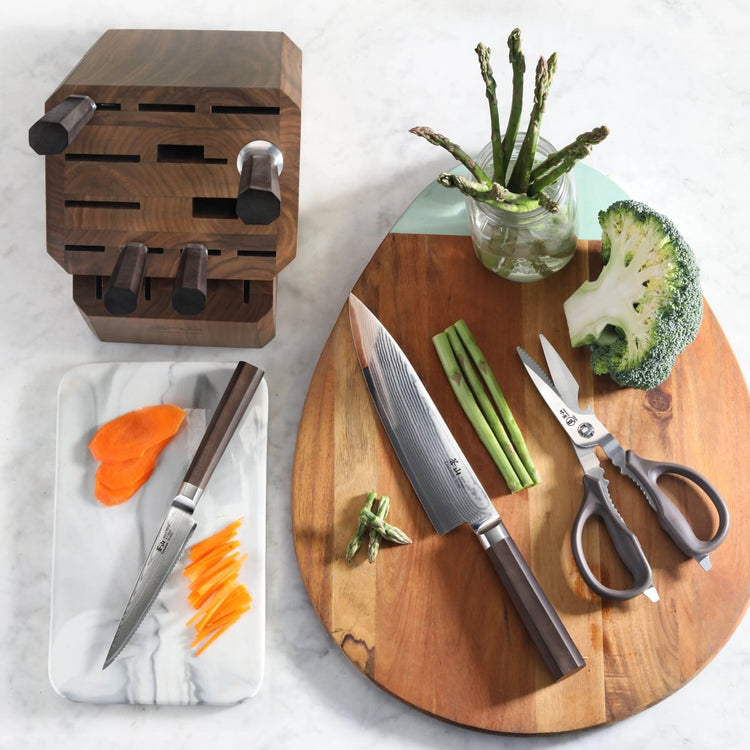 Wooden knife block with seven knives, a cutting board with a chef knife, scissors, asparagus, broccoli, and sliced carrots.