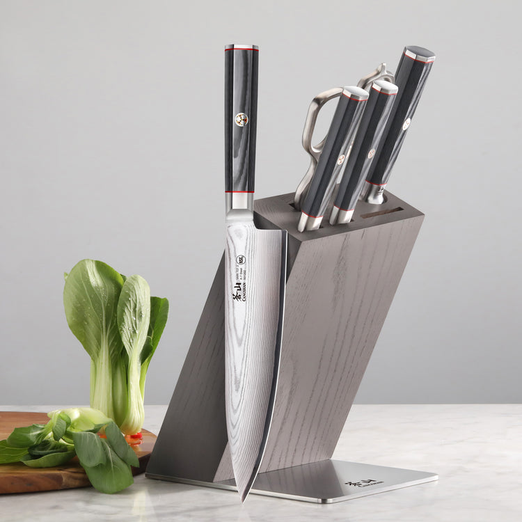 Six kitchen knives with black handles in a gray angled knife block on a marble surface next to leafy greens.