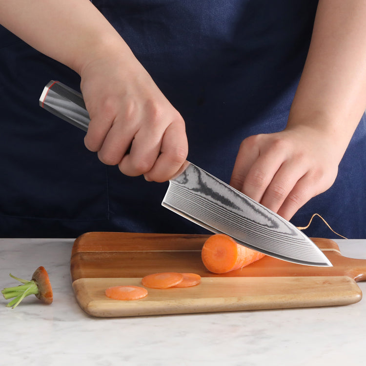 Hand holding a Damascus steel chef's knife slicing a carrot on a wooden cutting board.