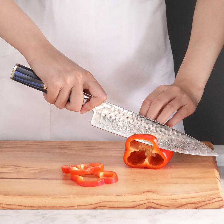 Hands holding a blue-handled knife slicing a red bell pepper on a wooden cutting board.