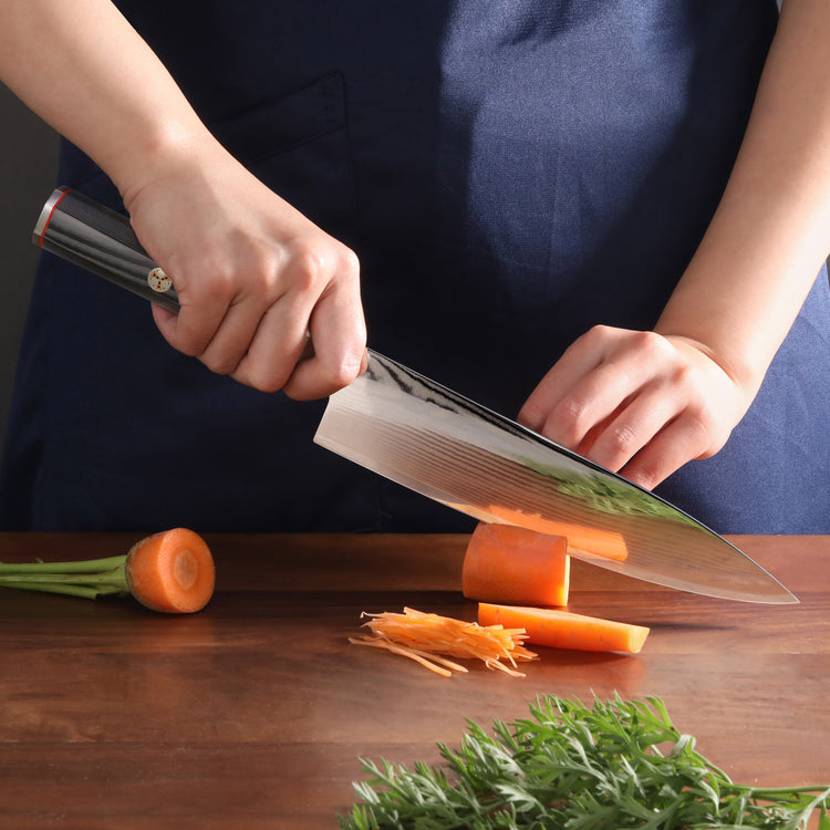 Hands holding a knife slicing a carrot on a wooden cutting board with carrot greens nearby.