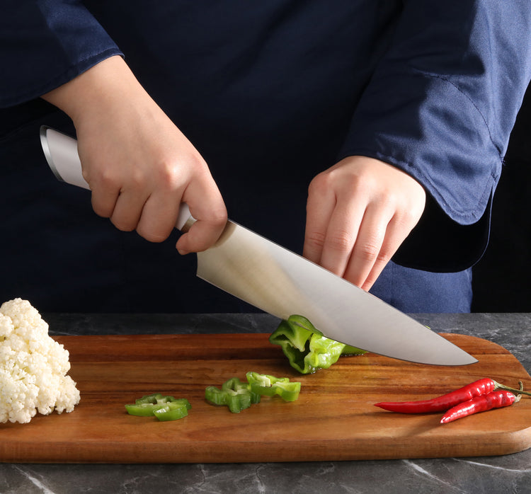 Hands slicing a green bell pepper on a wooden cutting board with a large chef's knife and red chili peppers nearby.