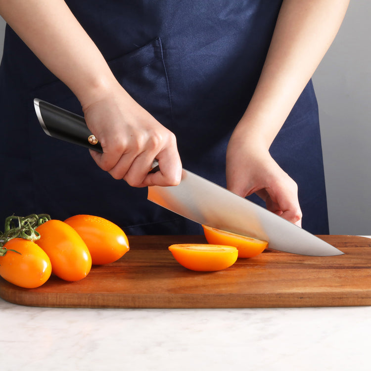 Hand holding a chef's knife cutting a yellow tomato on a wooden cutting board with more tomatoes nearby.