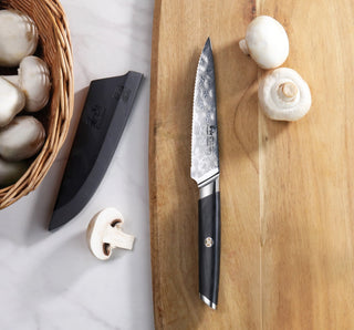 
                    
                      AIRA Series 5-Inch Serrated Utility Knife with 135-Layer Damax™ Steel on a wooden cutting board with mushrooms and a black
                    
                  