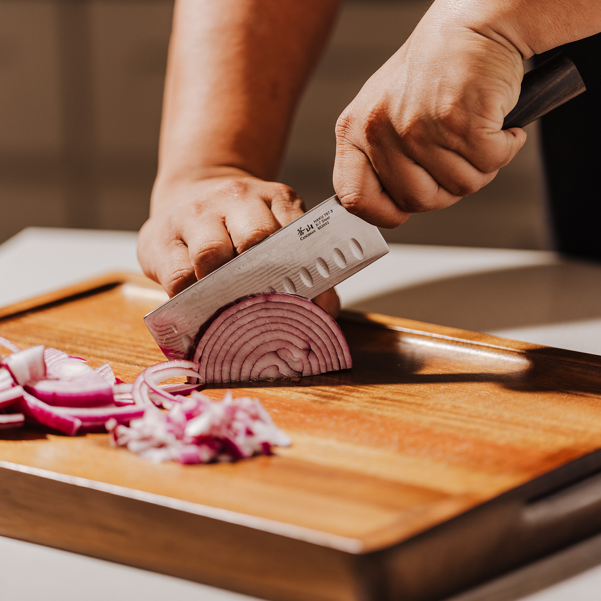 Hands slicing a red onion with a chef's knife on a wooden cutting board.
