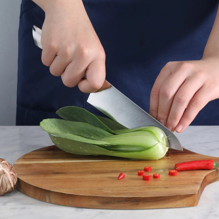 Hand holding a utility knife slicing green bok choy on a wooden cutting board with chopped red chili pieces nearby.