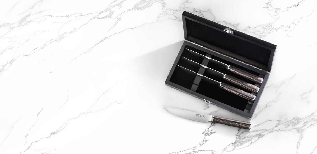 A set of four kitchen knives with dark wood handles and silver blades, shown in an open black presentation box on a white marble surface, with one knife placed outside the box.