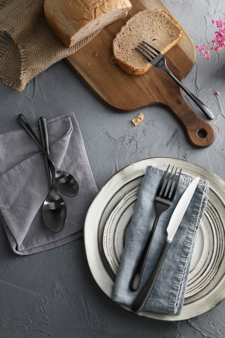 RAIN II Series black forged flatware set displayed on plates and napkins next to bread on a cutting board.