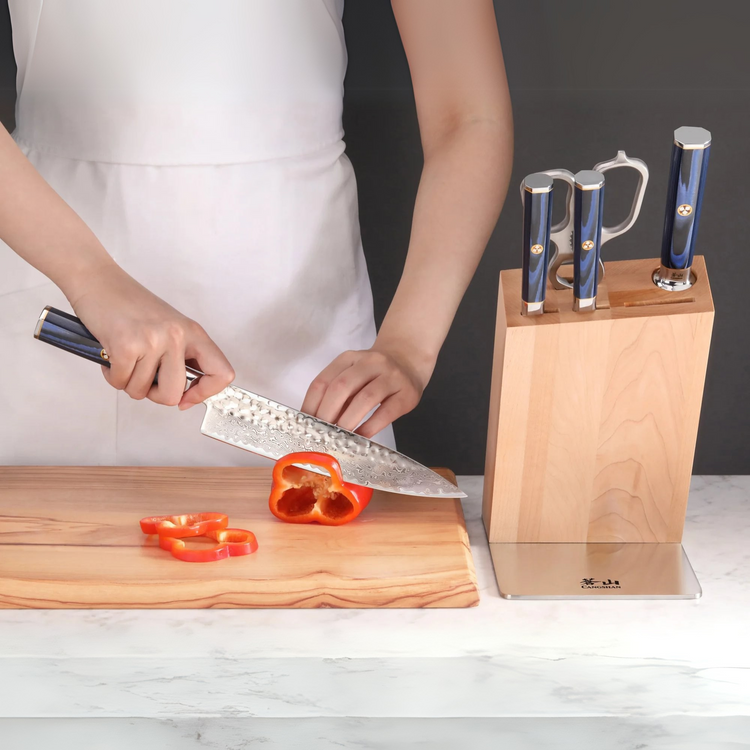 Person slicing a red bell pepper on a wooden cutting board next to a knife block with four knives.