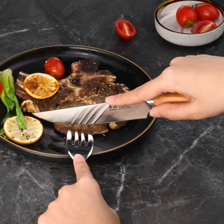 Hands using a steak knife with wooden handle to cut grilled meat on a black plate with lemon slices and vegetables.