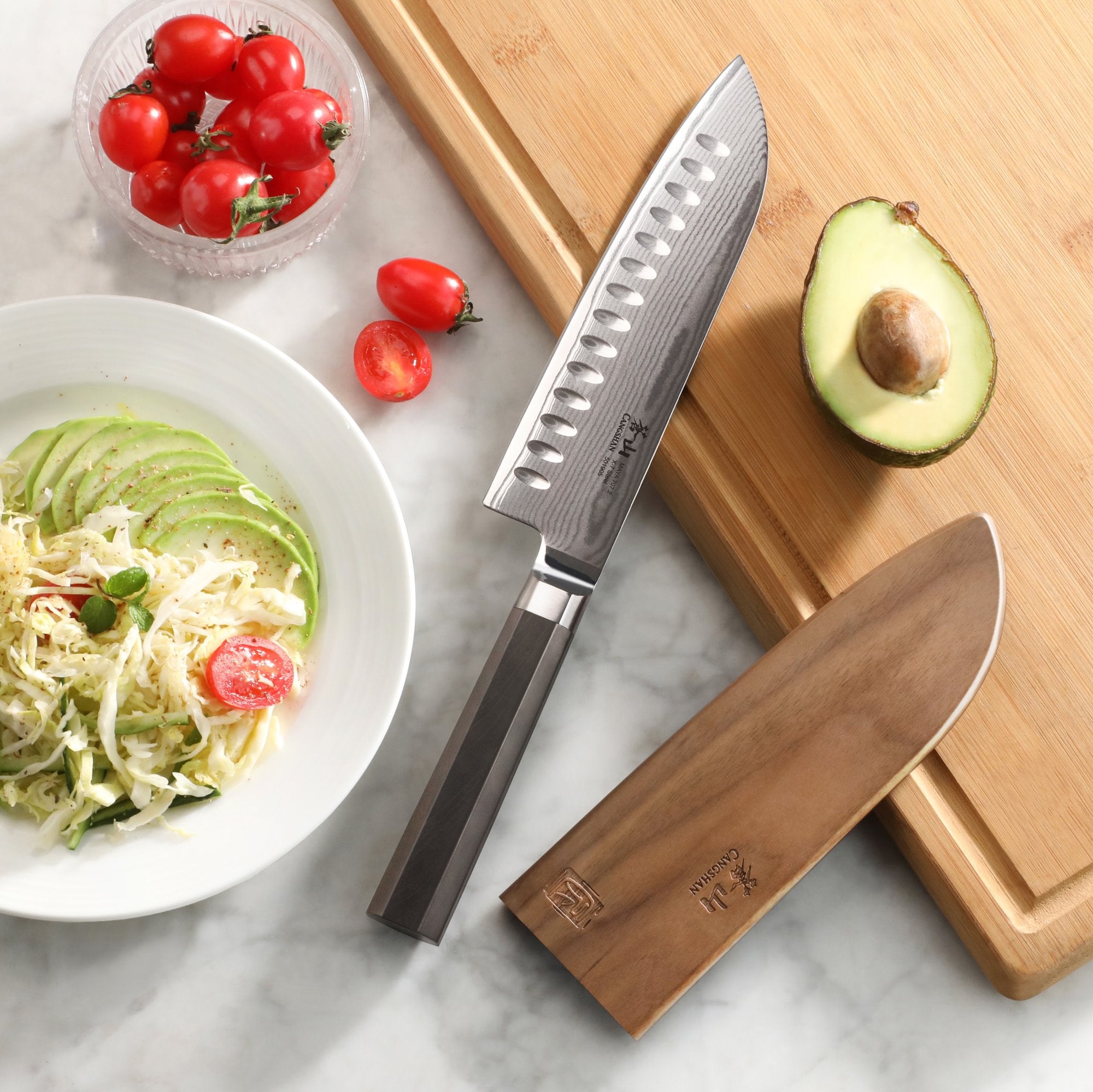 MAYA Series 7-inch Santoku Knife X-7 Damascus Steel with wooden sheath, avocado, and tomatoes on a cutting board.