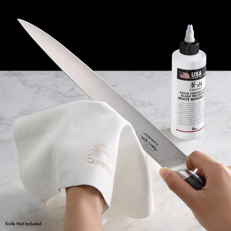 Hand polishing a large kitchen knife blade with a white cloth next to a bottle of blade protector on a marble surface.